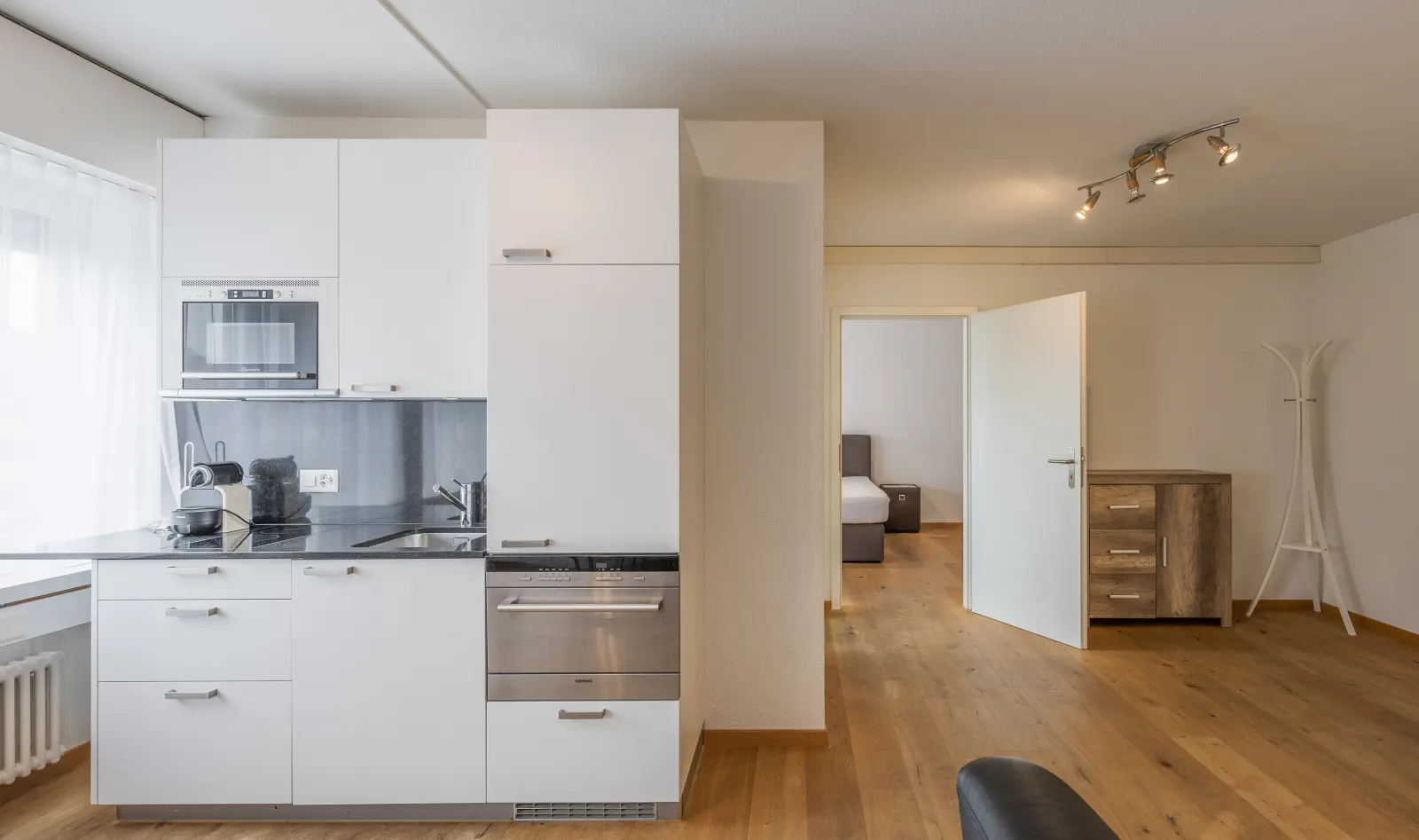 Weisse Einbauküche mit Backofen und Blick durch den Flur in ein Schlafzimmer